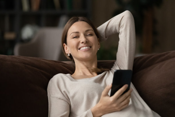 女人放松地坐在沙发上，拿着智能手机微笑着看着相机图片下载