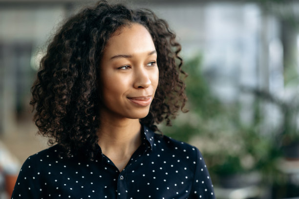 特写照片，一个漂亮的，沉思的深色皮肤的女孩，学生，经理，秘书，自由职业者或公司经理，站在办公室，穿着时髦的商务服装，望向别处，微笑图片下载
