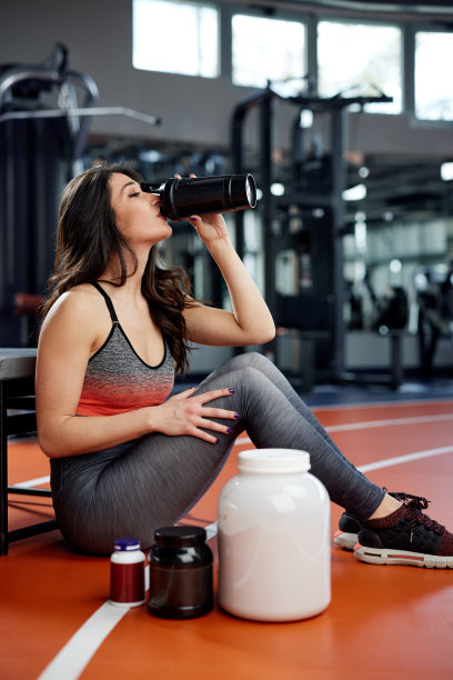 一个苗条的女运动员坐在健身房的地板上，周围都是补品，喝着蛋白质奶昔。图片下载