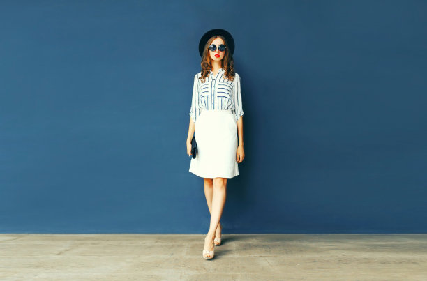 全长美丽的年轻女子穿着白色条纹衬衫，黑色圆帽，手提包手提包在深蓝色的背景图片下载