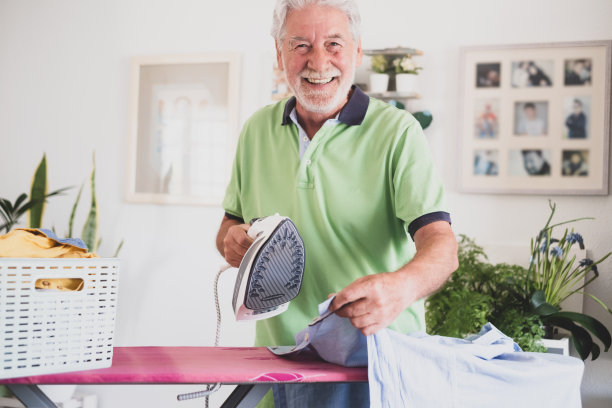 微笑着在熨衣板上熨烫衣服、手拿熨斗、看着镜头的老年男子的肖像图片下载