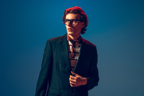 时髦的年轻人肖像在官方夹克和黑色眼镜，摆姿势，看远离孤立的蓝色工作室背景在霓虹灯图片下载