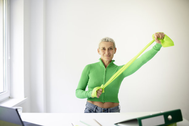 中年女商人站在白色高桌旁，用绿色的弹力带或体操带做伸展运动图片下载