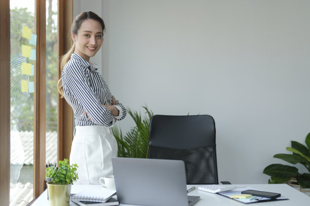 自信的千禧一代女商人双手交叉站在办公室窗户旁，对着镜头微笑。图片下载
