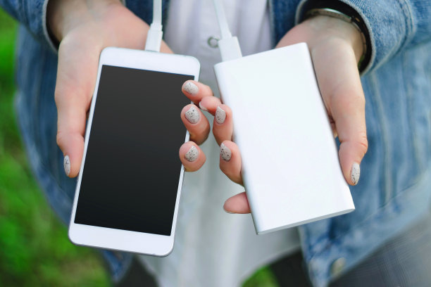 绿色草地上的女孩拿着充电宝和智能手机。女孩用充电宝给智能手机充电。图片下载