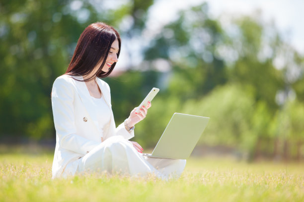 美丽的商业女性在公园户外工作。年轻可爱的女士穿着白色西装坐在绿色的草地上，带着笔记本电脑，打电话。图片下载