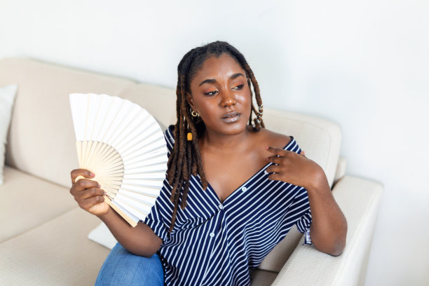 压力烦恼的非洲妇女用着挥舞的电扇遭受过热，夏天热健康荷尔蒙问题，没有空调坐在家里的沙发上感到疲惫脱水中暑的概念图片下载
