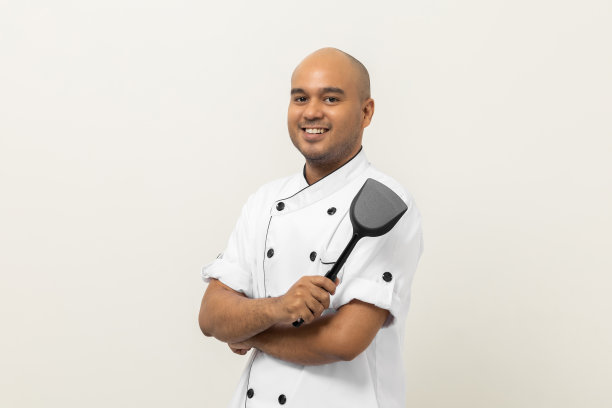 微笑的年轻英俊的亚洲男子厨师在制服站与双臂交叉持有特纳厨房用具在孤立的背景。厨师职业厨师在厨房、餐厅和酒店工作。图片下载