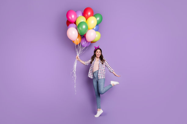 全身尺寸照片开朗快乐的生日女孩保持桩气球穿着牛仔裤衬衫孤立在生动的紫色背景图片下载