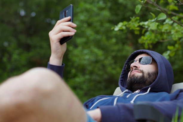 带着墨镜的大胡子白人男子在露营椅上用智能手机休息。娱乐的概念图片下载