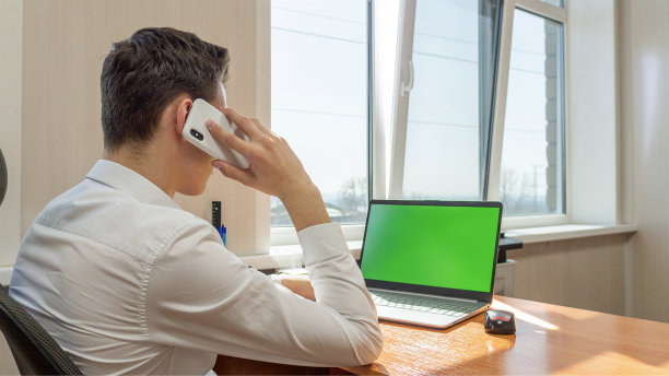 一个年轻人坐在办公室的绿色屏幕的电脑前打电话图片下载