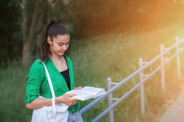 年轻的女学生在城市公园里倚着金属栏杆看书。图片下载
