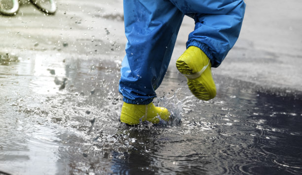 孩子们的脚穿着胶靴和不湿的裤子。一个孩子跳进水坑里溅起水花图片下载