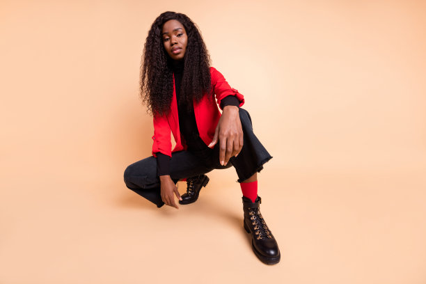 低角度查看全身照片的非洲美国惊人的女士坐穿靴子红色运动上衣孤立在米色背景图片下载