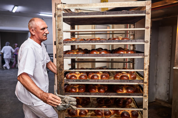在一家面包店，穿着制服的专业面包师从工业烤箱里拿出一辆装着刚烤好的面包的推车图片下载