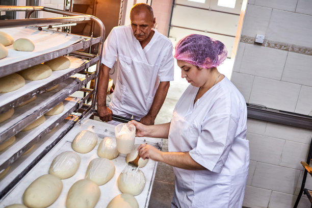 专业的面包师穿着制服在面包房的同事背景上涂抹黄油面包面团准备烘焙图片下载