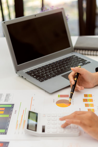 商人拿着笔用财务图工作，笔记本电脑用计算器在办公室分析财务数据，垂直图像。图片下载