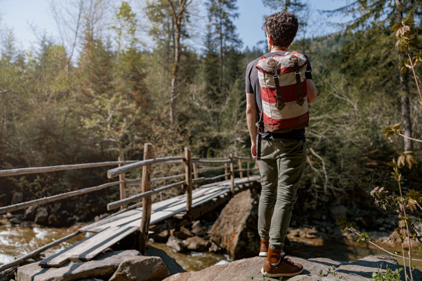 背双肩包的男子站在山上的岩石上，在河上的木桥上。图片下载