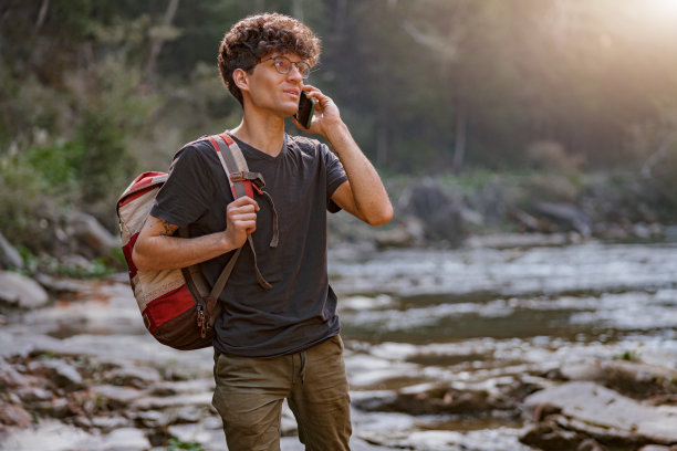 年轻的男性旅行者在山上走在河边，用手机聊天。徒步旅行。图片下载