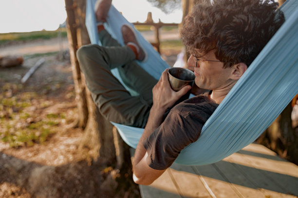 年轻英俊的男游客身后躺在吊床上，手里拿着保温杯。图片下载