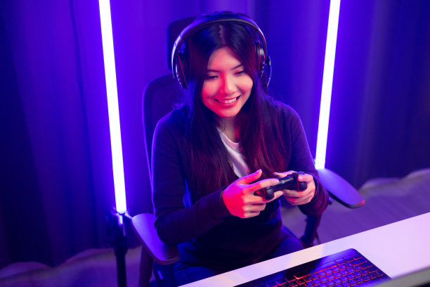 玩视频游戏。年轻的亚洲漂亮女人坐在椅子上握着操纵杆在客厅。快乐女职业主播中国人戴着耳机在黑暗的房间里玩游戏霓虹灯。图片下载