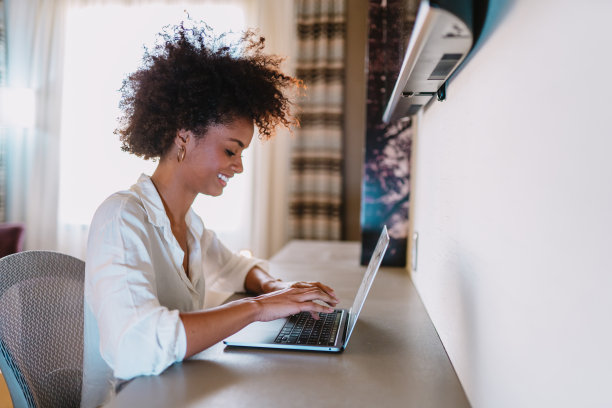 专注于工作。自信的年轻拉丁女子在聪明的休闲服装在笔记本电脑上工作，而坐在酒店的卧室。图片下载