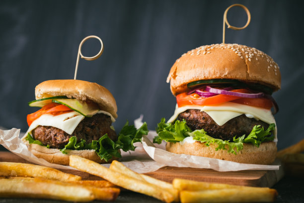 大牛肉饼汉堡配奶酪，西红柿，紫色洋葱，黄瓜和生菜，旁边是炸薯条，黑色的木制支架，黑色的背景图片下载