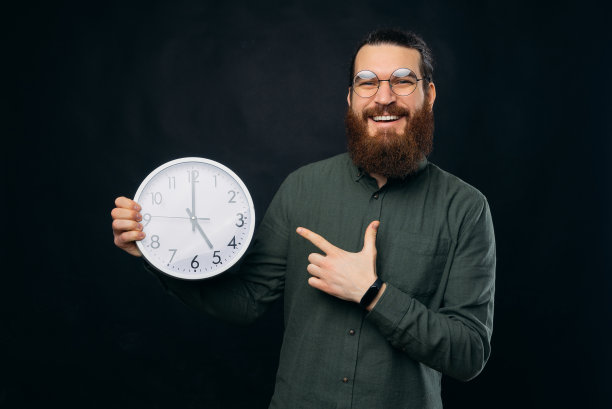 年轻的胡子男子戴着圆形眼镜指着黑色背景上的白色时钟的照片图片下载