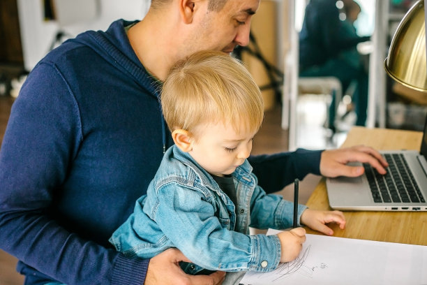 父亲抱着年幼的儿子在家里用笔记本电脑工作。远程工作的概念图片下载