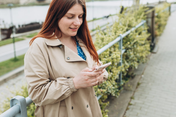 美丽快乐的红发女人在户外用智能手机微笑。城市生活方式的概念。走在城市街道上的女孩查看她的智能手机。春天的时间图片下载