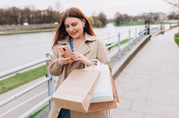 美丽的女人拿着购物纸袋，用着智能手机，在户外微笑。城市生活方式的概念。图片下载