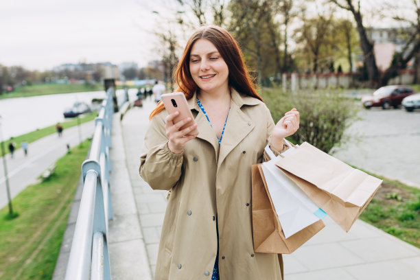 美丽快乐的红发女子手持购物纸袋，使用智能手机，在户外微笑。城市生活方式的概念。图片下载