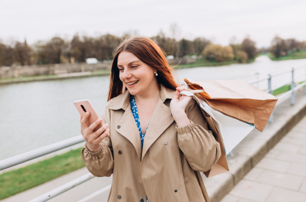 美丽的女人拿着购物纸袋，用着智能手机，在户外微笑。城市生活方式的概念。图片下载