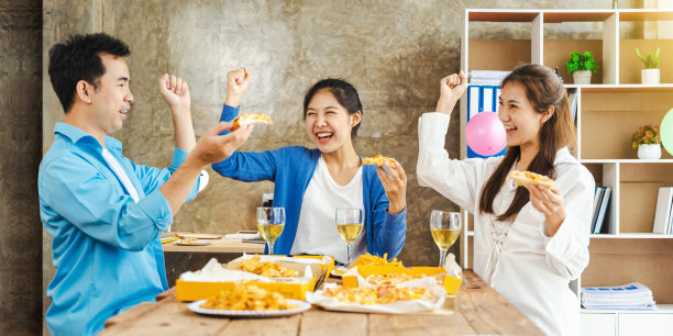 亚洲人一起坐在办公室里吃披萨。上班族们坐在办公室的桌子旁，高兴地举手吃午饭。图片下载