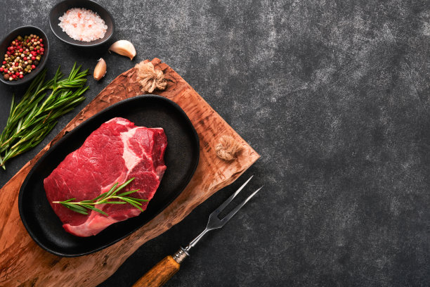 牛排。新鲜生牛肋眼牛排，叉上迷迭香，盐和胡椒粉，放在烤盘上，底色为深灰色或黑色。商店广告的想法。前视图。设计理念的模型。图片下载