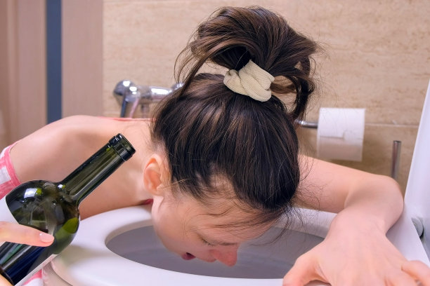 醉醺醺的女人正吐在马桶里，手里拿着酒瓶坐在家里的地板上，早上宿醉，侧看。图片下载