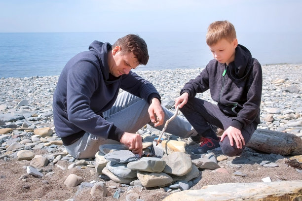 爸爸和儿子在海边手工制作的石头火盆上点燃篝火中的煤块。家庭的周末。图片下载