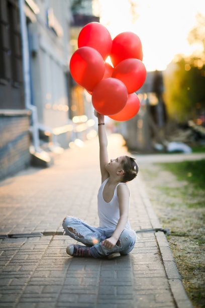 一个穿着牛仔裤和白t恤的女孩坐在人行道上，手里拿着一堆红气球，这是Z一代，孤独的节日图片下载