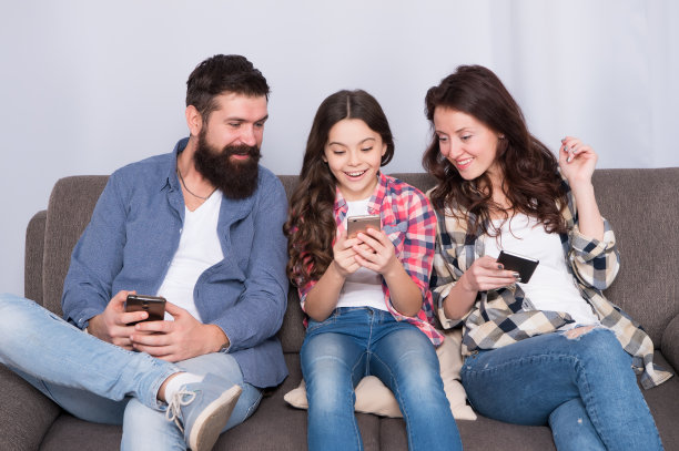 玩游戏的应用程序。家庭网络冲浪。家庭休闲。网上的家庭。妈妈，爸爸和女儿在沙发上休息。家人在网上度过周末。孩子小女孩和父母一起使用智能手机图片下载