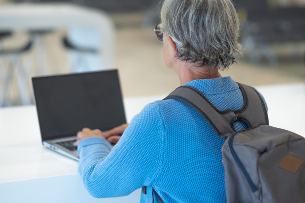 后视图成熟的灰色头发的女人背双肩包坐在机场等待航班起飞，同时使用笔记本电脑在白色的桌子上图片下载