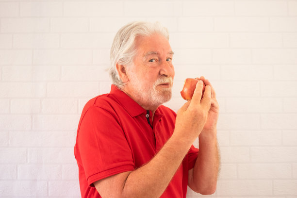 在白色背景上穿着红色马球衫的滑稽老人，嘴里咬着一口苹果，手里拿着一口被咬过的红苹果图片下载