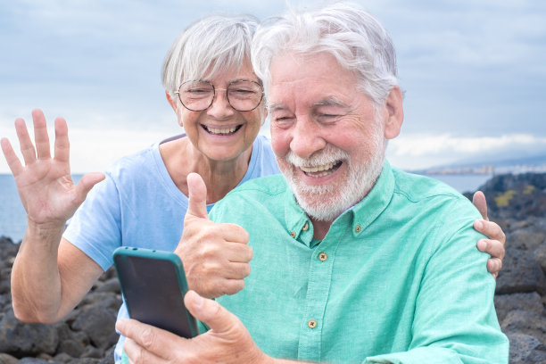 快乐的白人老年夫妇坐在户外在海上视频电话与手机手势积极的迹象。两位老人在使用手机的同时享受着自由和放松。地平线在水图片下载