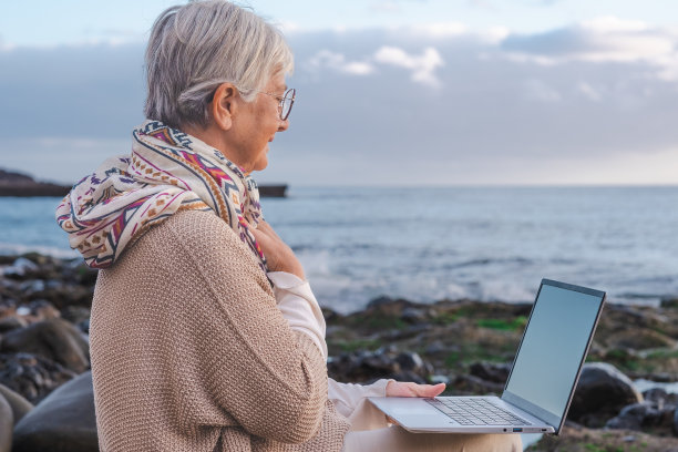 年老的活跃的女人坐在卵石滩上使用笔记本电脑远程工作。成熟的女性在海上享受日落，膝上拿着笔记本电脑，水面上的地平线图片下载