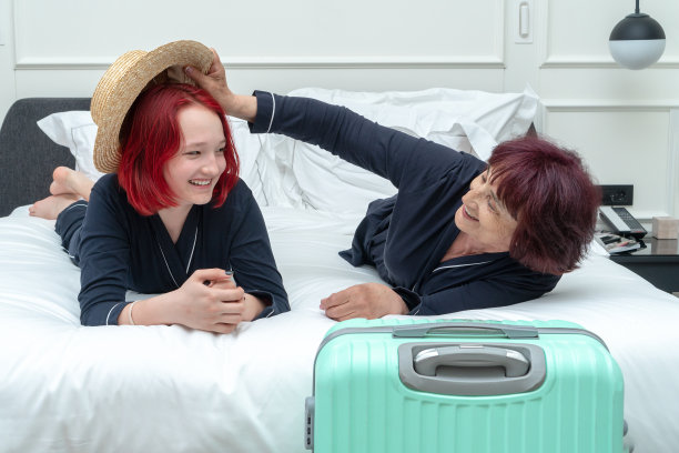 在一个时髦的酒店房间里，一个穿着睡衣、戴着帽子的年长女性和一个红头发的少女躺在床上，背景是一个行李箱。垂直的照片。与家人在时尚酒店度假的概念。的生活方式。坦诚的家庭。图片下载