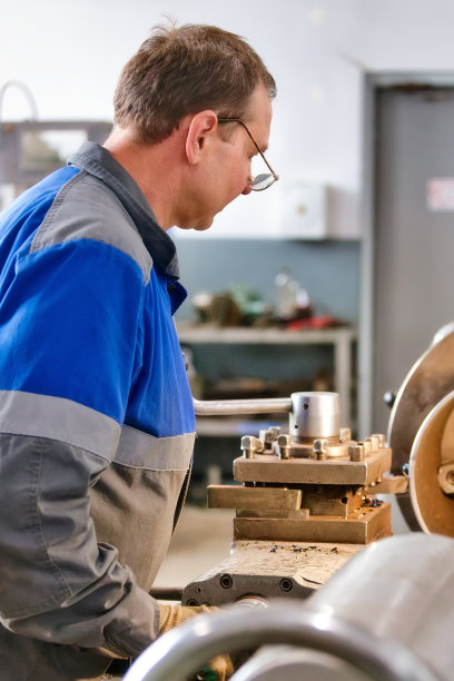 50-55岁穿着工装的老车工在生产车间的车床旁工作。金属制品加工。图片下载