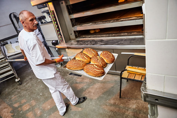 在一家面包店，穿着制服的专业面包师从工业烤箱里拿出一辆装着刚烤好的面包的推车图片下载