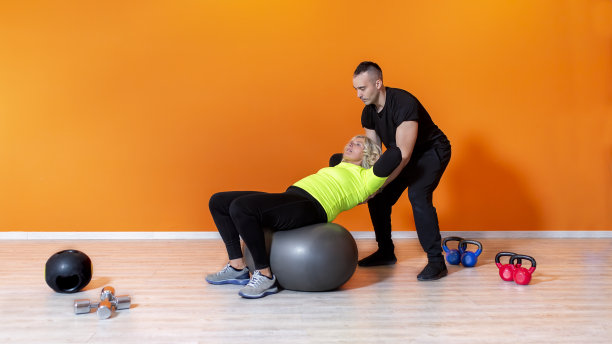 年长健身妇女做锻炼坐在一个球在健身房孤立的橙色背景，后面跟着一个私人教练。成年人室内健身训练。长者康复概念图片下载
