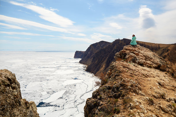 春天的贝加尔湖。一个女人坐在岩石上欣赏美丽的风景。白色的浮冰在水面上融化。图片下载