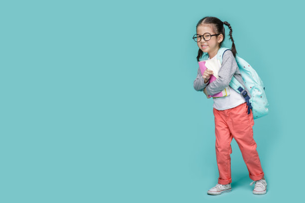 美丽微笑的亚洲小女孩戴着眼镜，拿着书与书包回到学校，在工作室的空白空间拍摄孤立的彩色蓝色背景，学校的教育理念素材图片