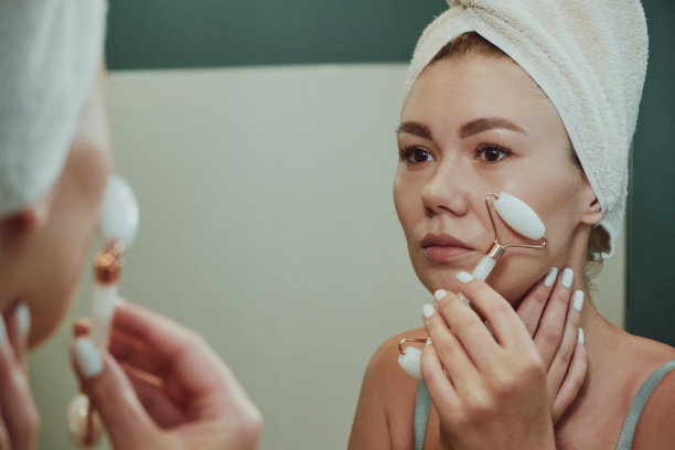 特写妇女使用玉石面部按摩辊在浴室的镜子看脸图片下载